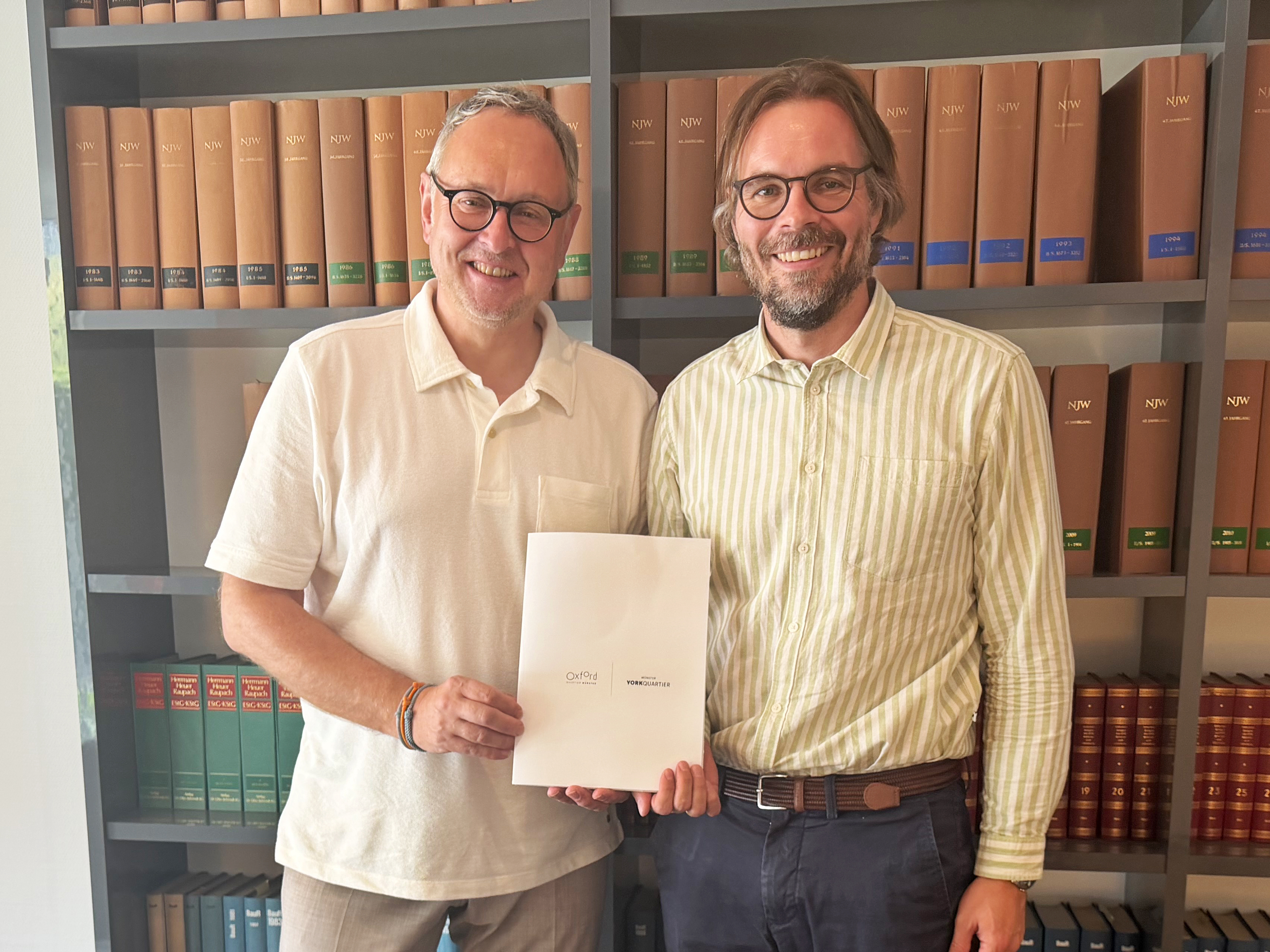 Zwei Männer stehen nebeneinander, lächeln in die Kamera und halten dabei ein Blatt Papier in den Händen
