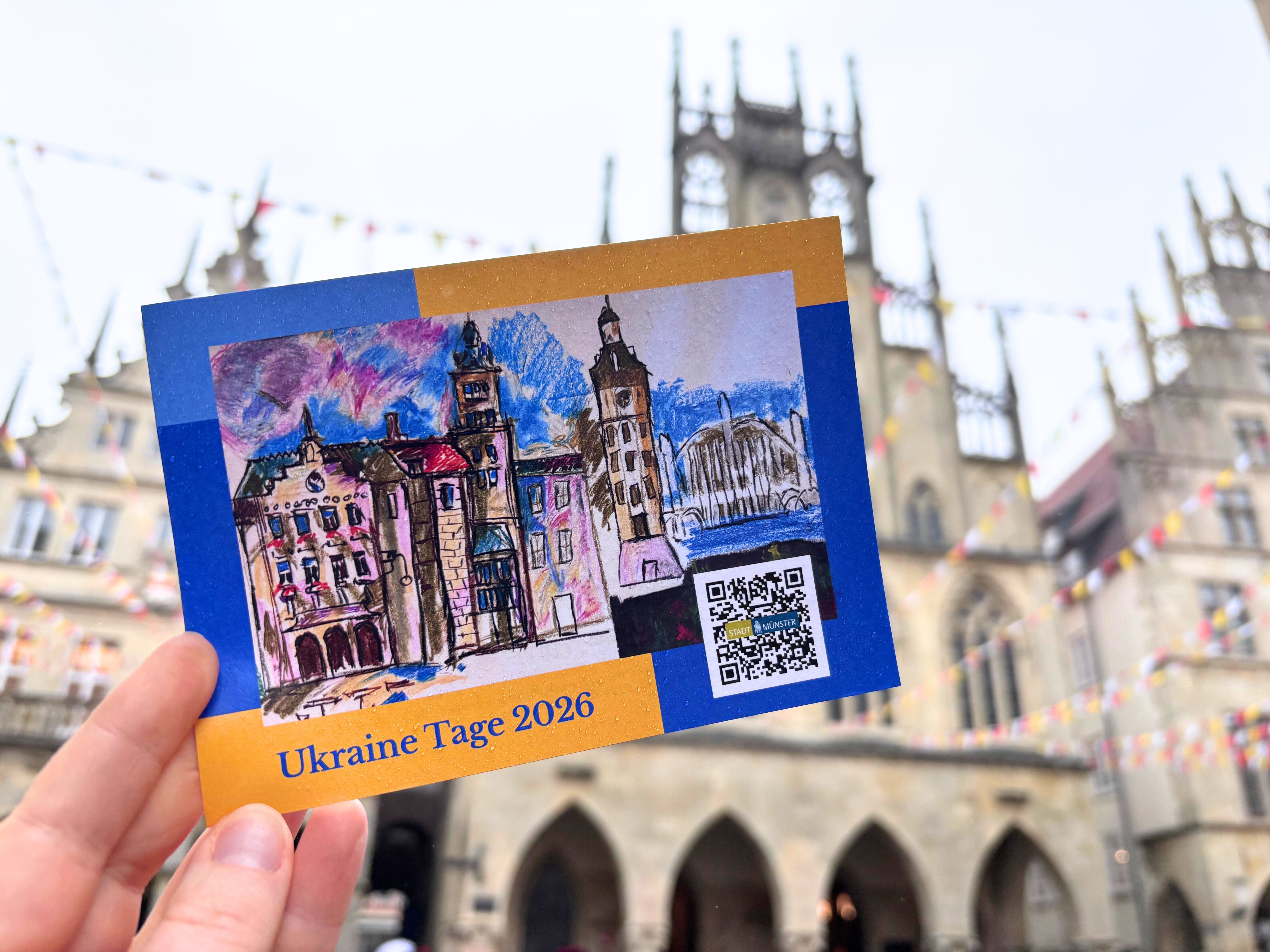 Eine Hand hält eine Postkarte zu der Veranstaltungsreihe Ukraine-Tage hoch. Im Hintergrund ist das Rathaus am Prinzipalmarkt unscharf zu erkennen. 