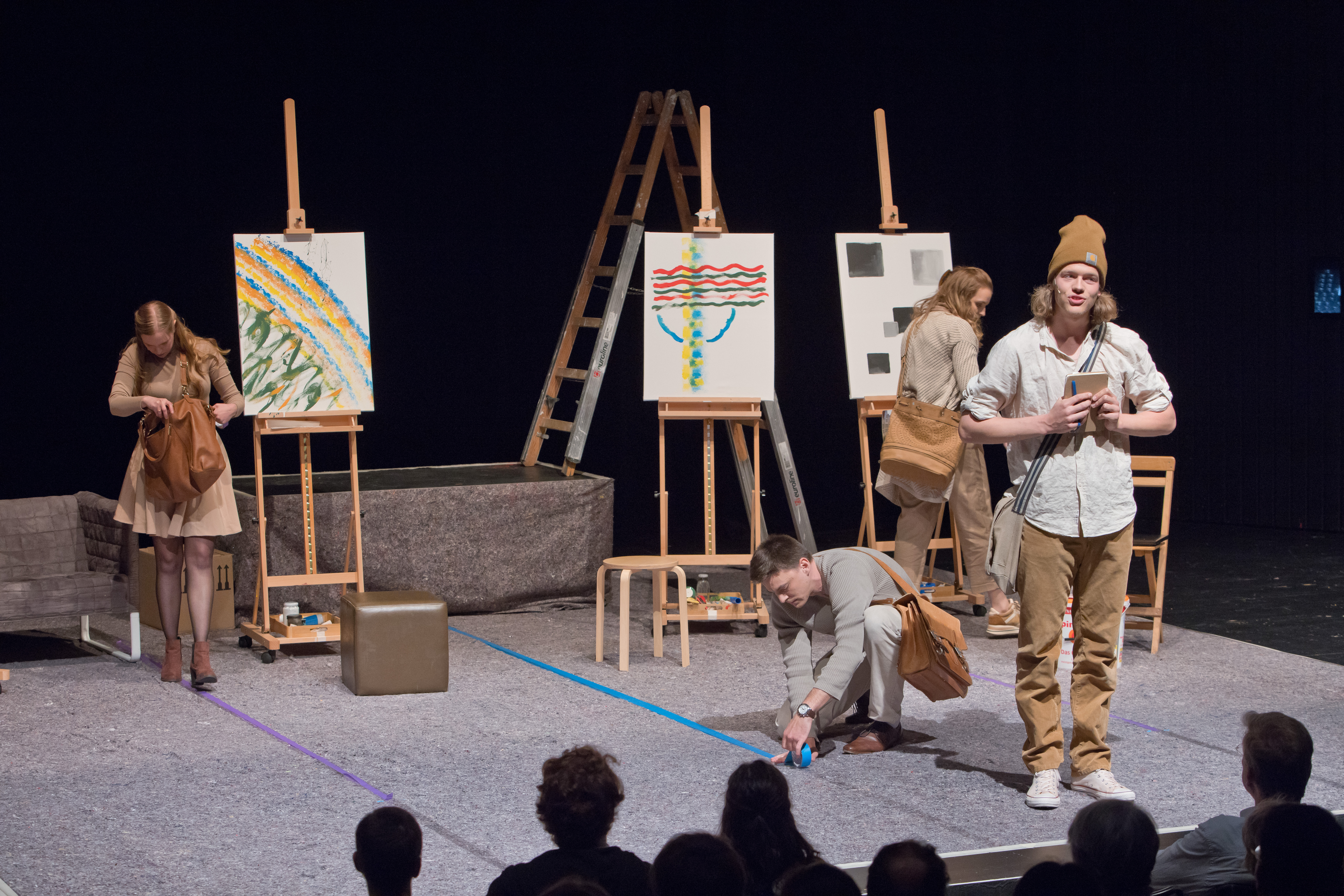 Vier Personen stehen verstreut auf einer Bühne. Im Hintergrund sind Staffeleien mit bemalten Leinwänden.