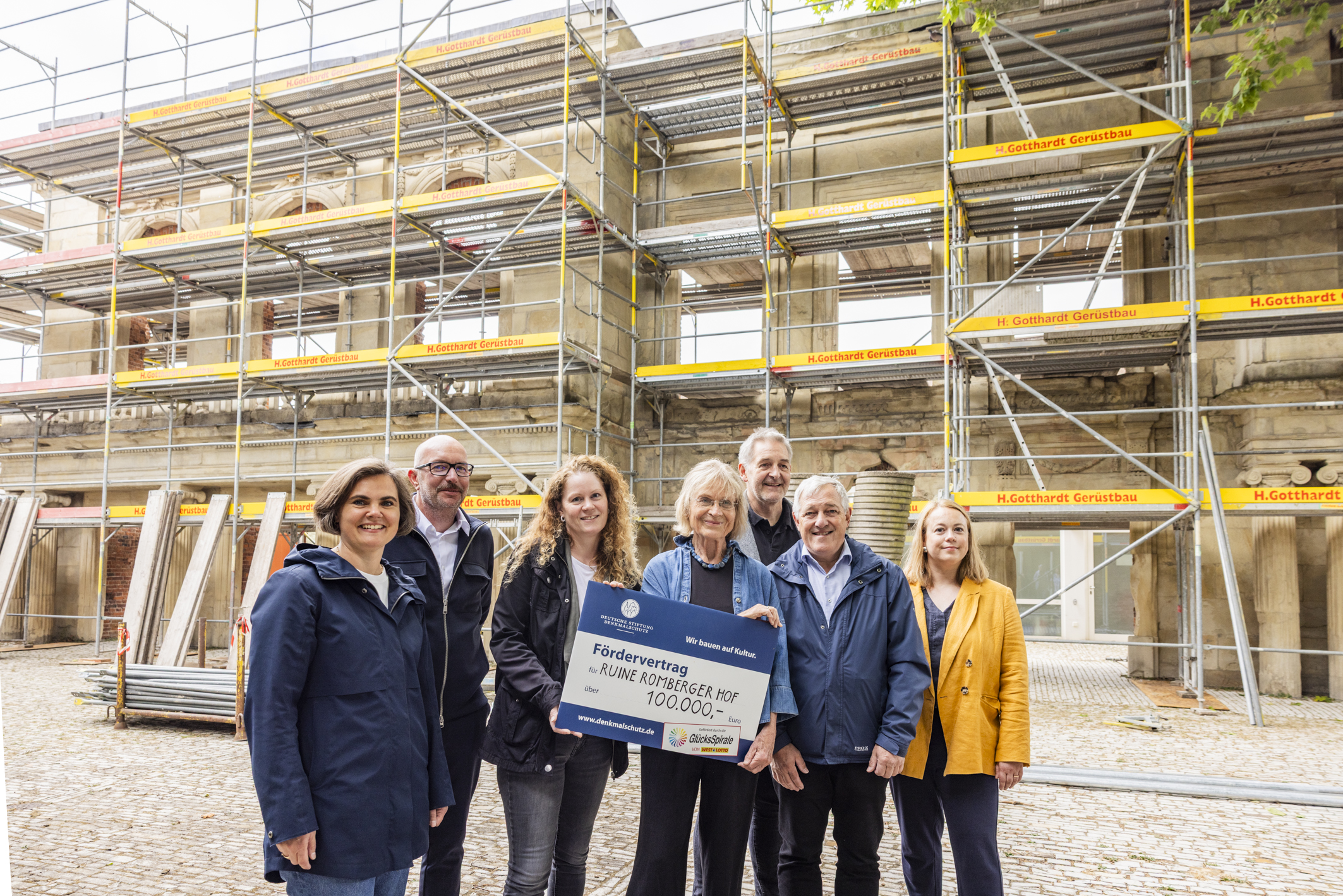 Der Fördervertrag in Höhe wird übergeben: Im Vordergrund die Beteiligten der Denkmalbehörde, Amt für Immobilienmanagements und der Deutschen Stiftung Denkmalschutz sowie des Theaters und von WestLotto. Im Hintergrund die Ruine mit Baugerüst.