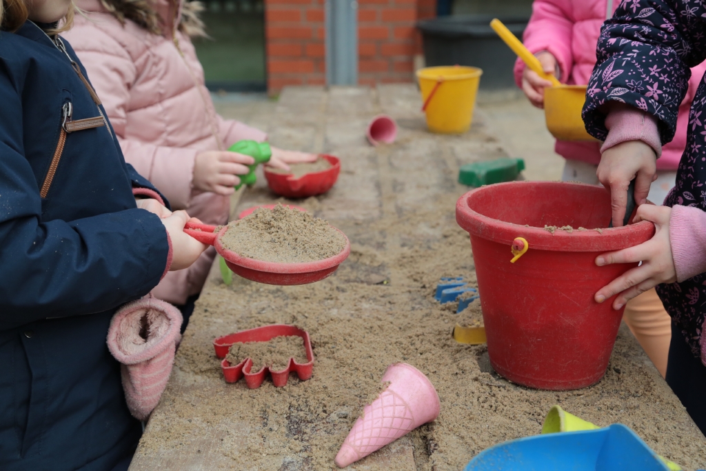 Mehrere Kinder spielen mit Sand auf einem Tisch. Sie halten Förmchen und Eimer. Es sind nur ihre Hände zu sehen. 