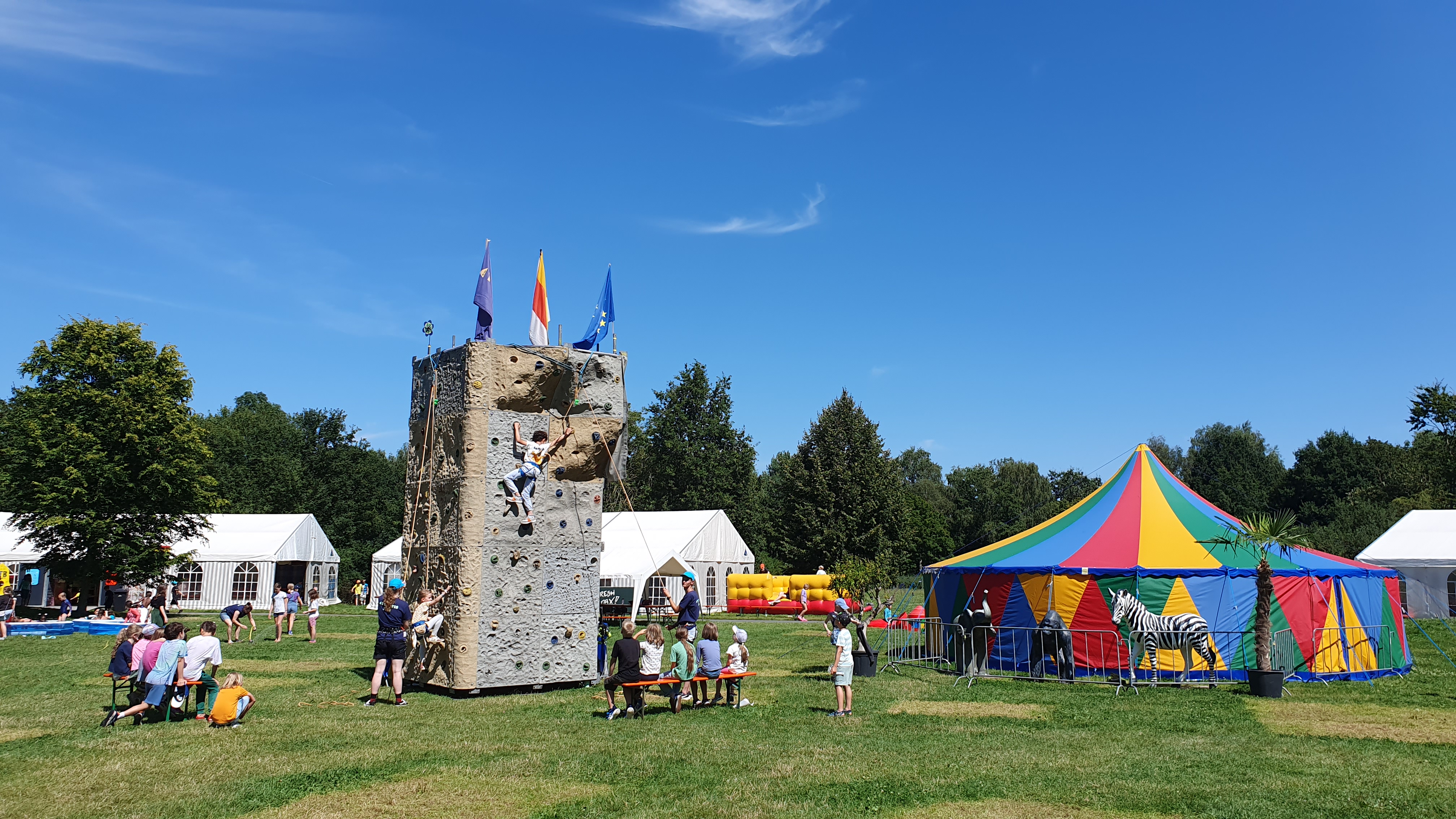 Eine große Wiese mit einem bunten Zirkuszelt und einem Kletterturm darauf.