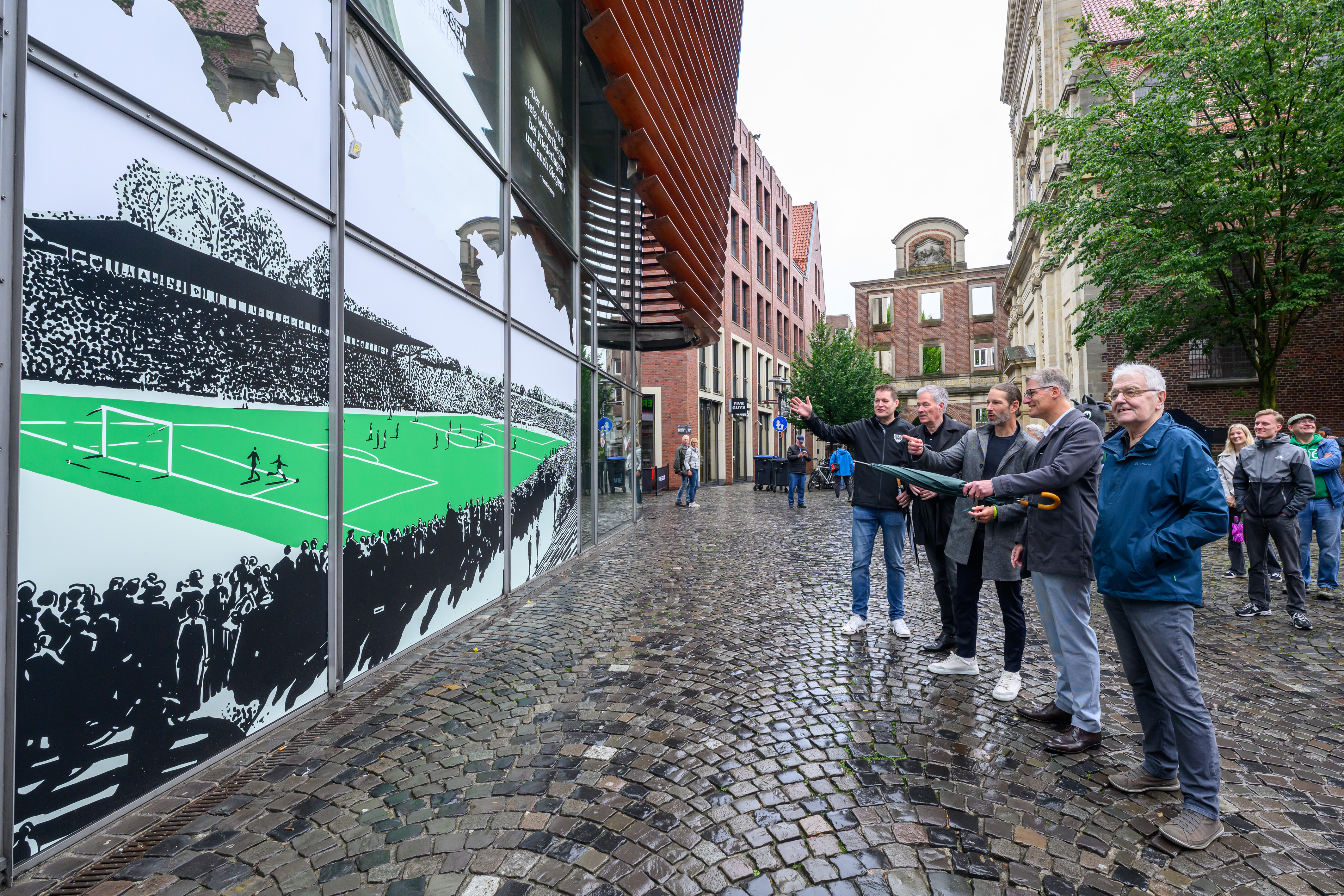 Fünf Männer schauen auf eine Illustration des Preußenstadions, aufgebracht auf einer Glasfassade