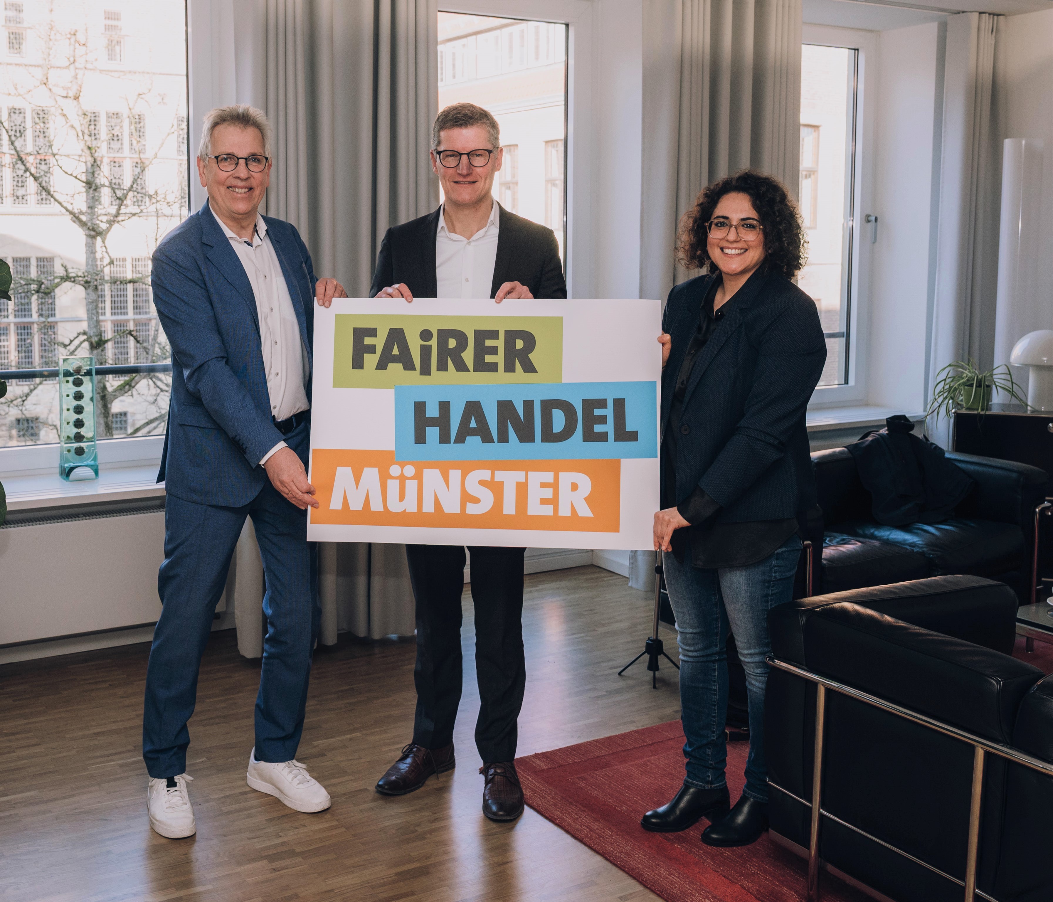 Drei Personen halten ein Plakat mit der Aufschrift "Fairer Handel Münster" in den Händen