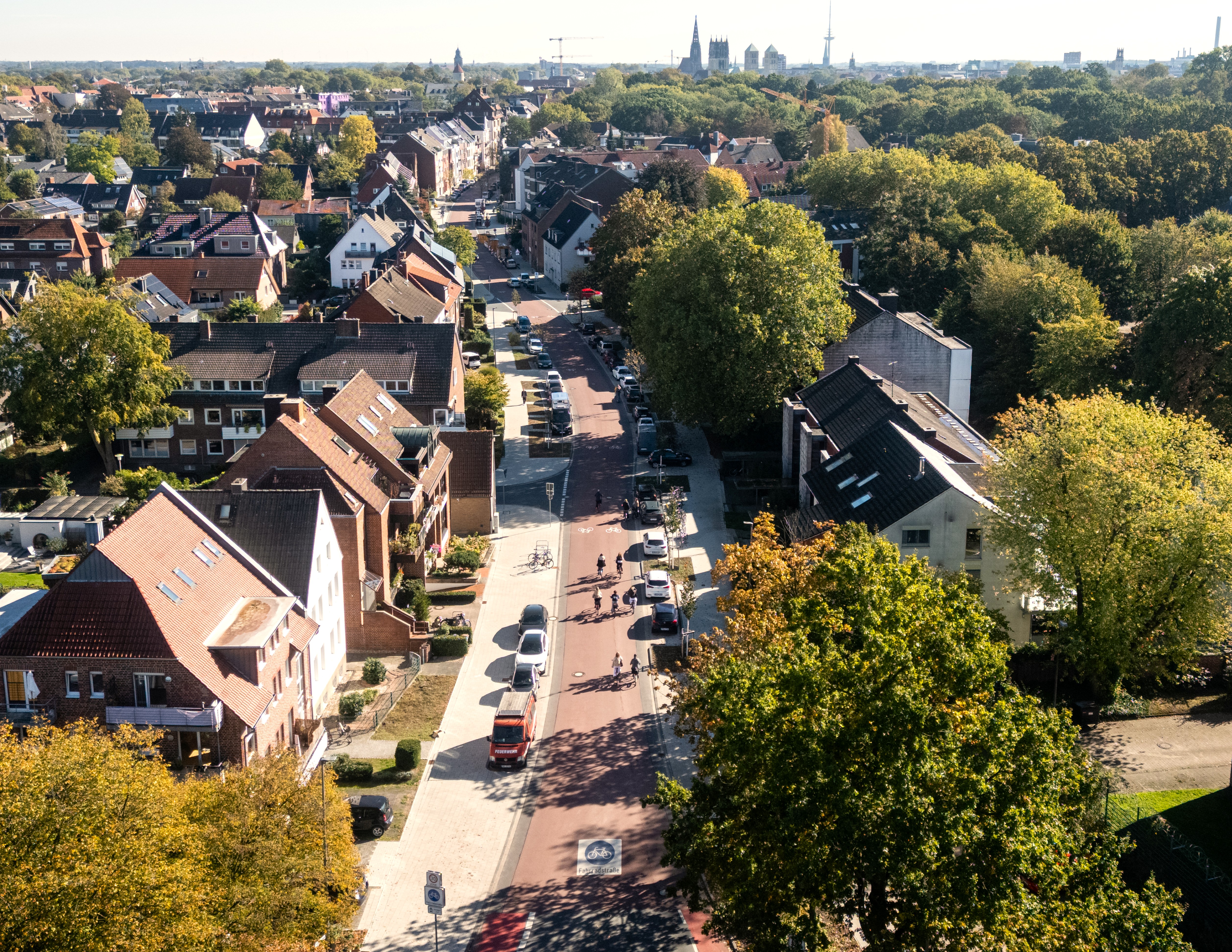 Luftbild auf die Wilhelmstraße mit roter Fahrbahn, Radfahrenden, am Straßenrand parkenden Autos, einigen Häusern und Bäumen. Im Hintergrund ist Münsters Skyline mit Lambertik- und Überwasserkirche, Dom und Fernsehturm.