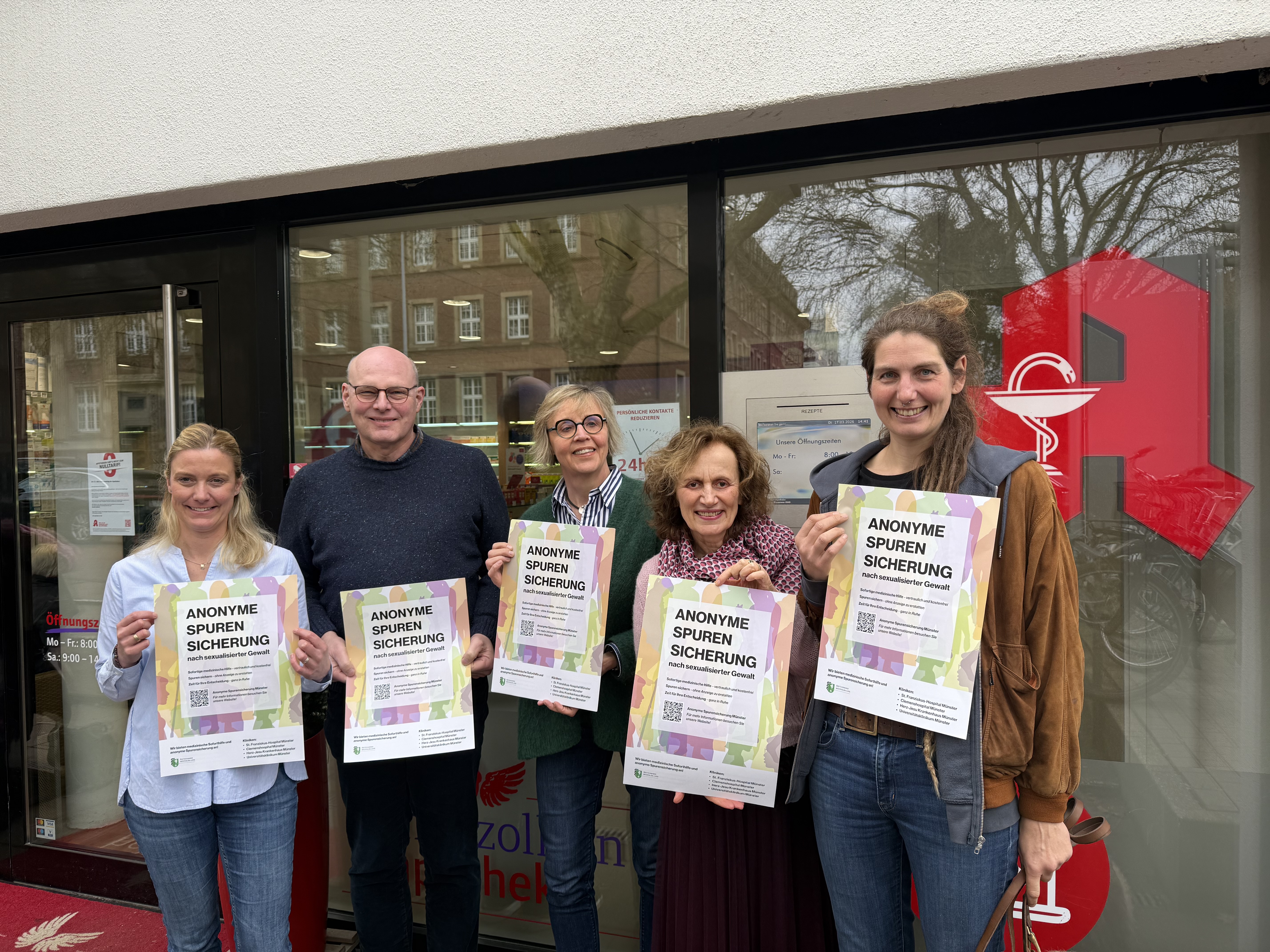 Fünf Personen stehen vor einer Apotheke und halten Plakate hoch