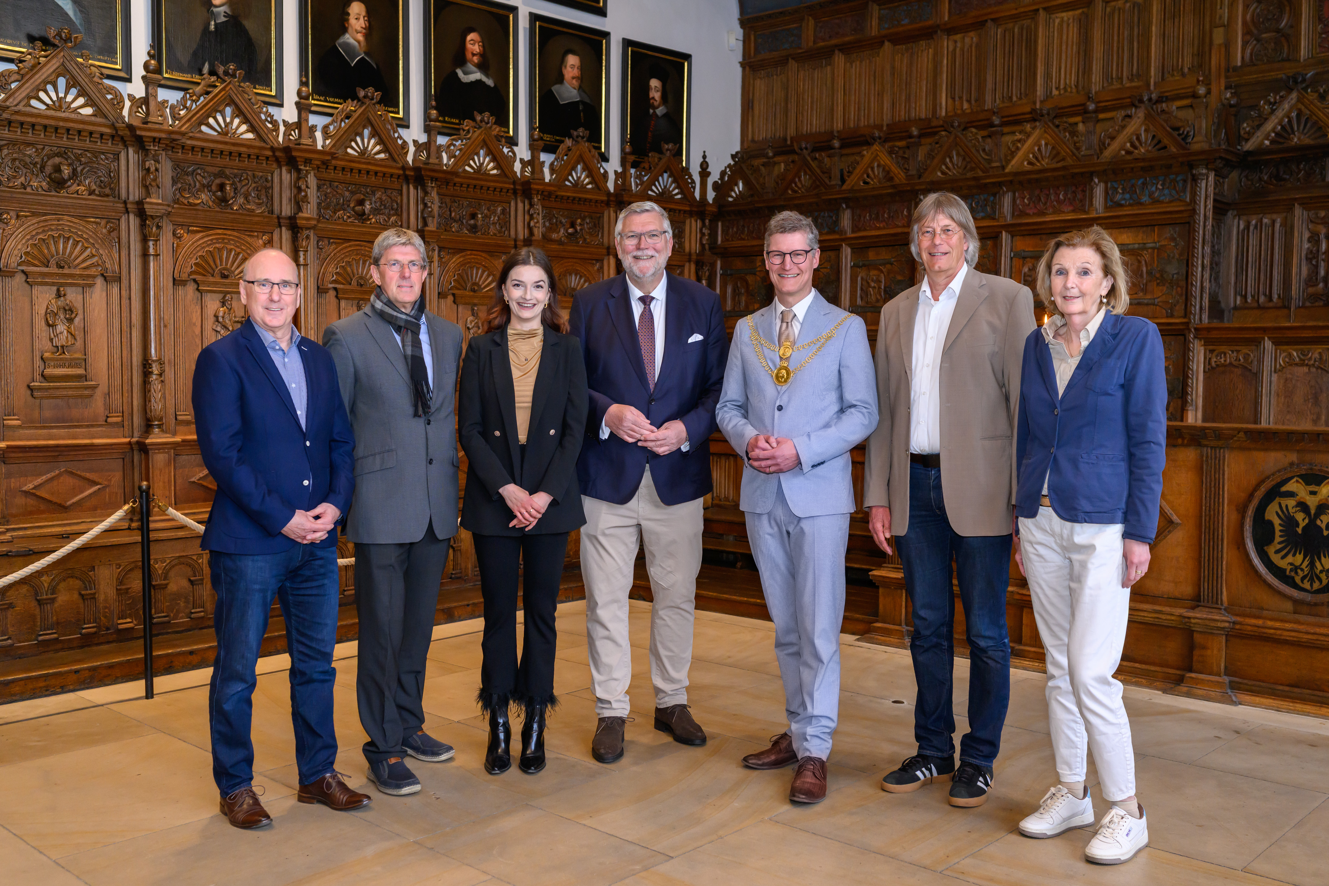 Sieben Personen stehen im holzvertäfelten Friedenssaal und lächeln in die Kamera