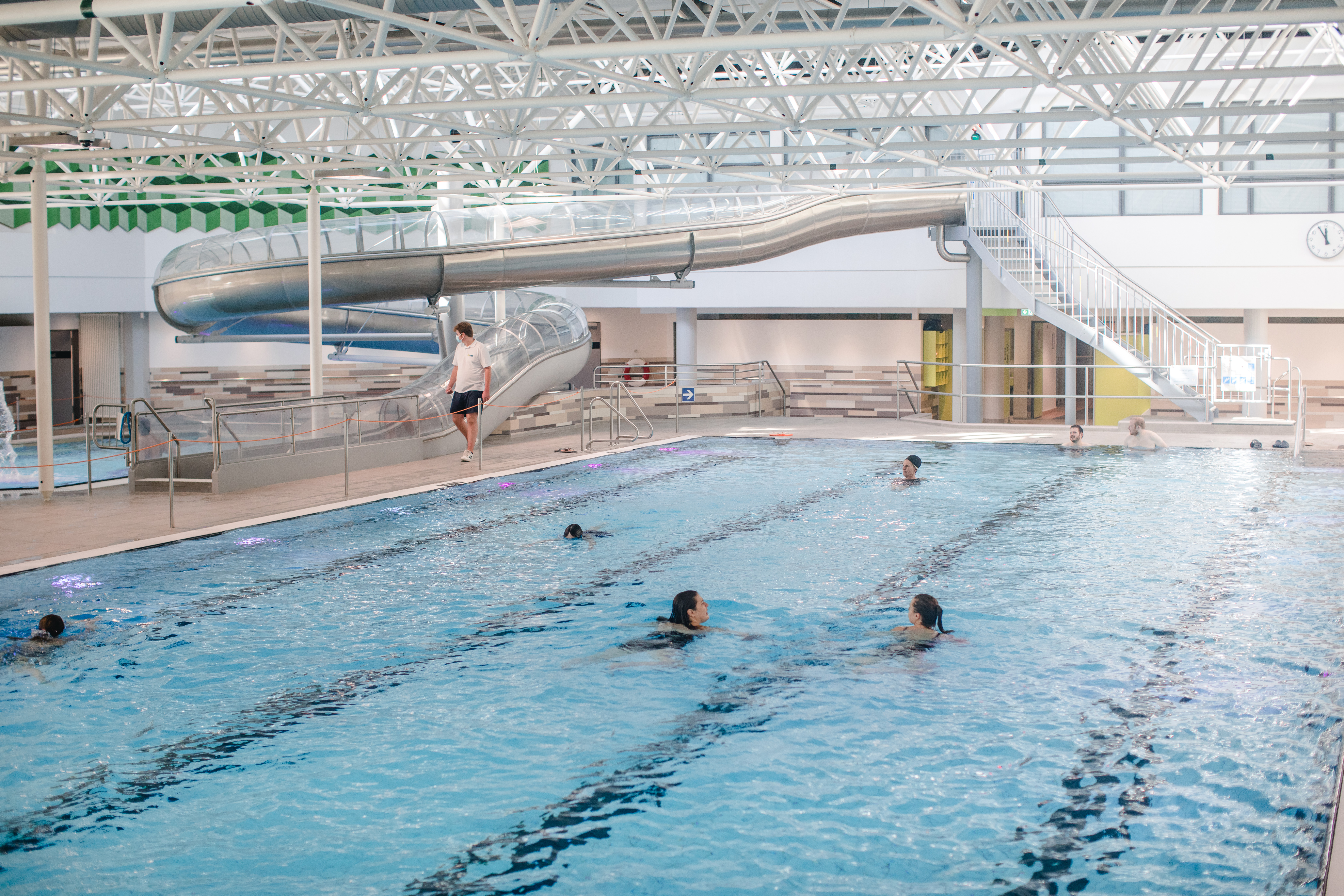 Blick auf das Schwimmerbecken im Hallenbad Kinderhaus. Ein paar Menschen sind im Wasser, im Hintergrund ist die Rutsche.