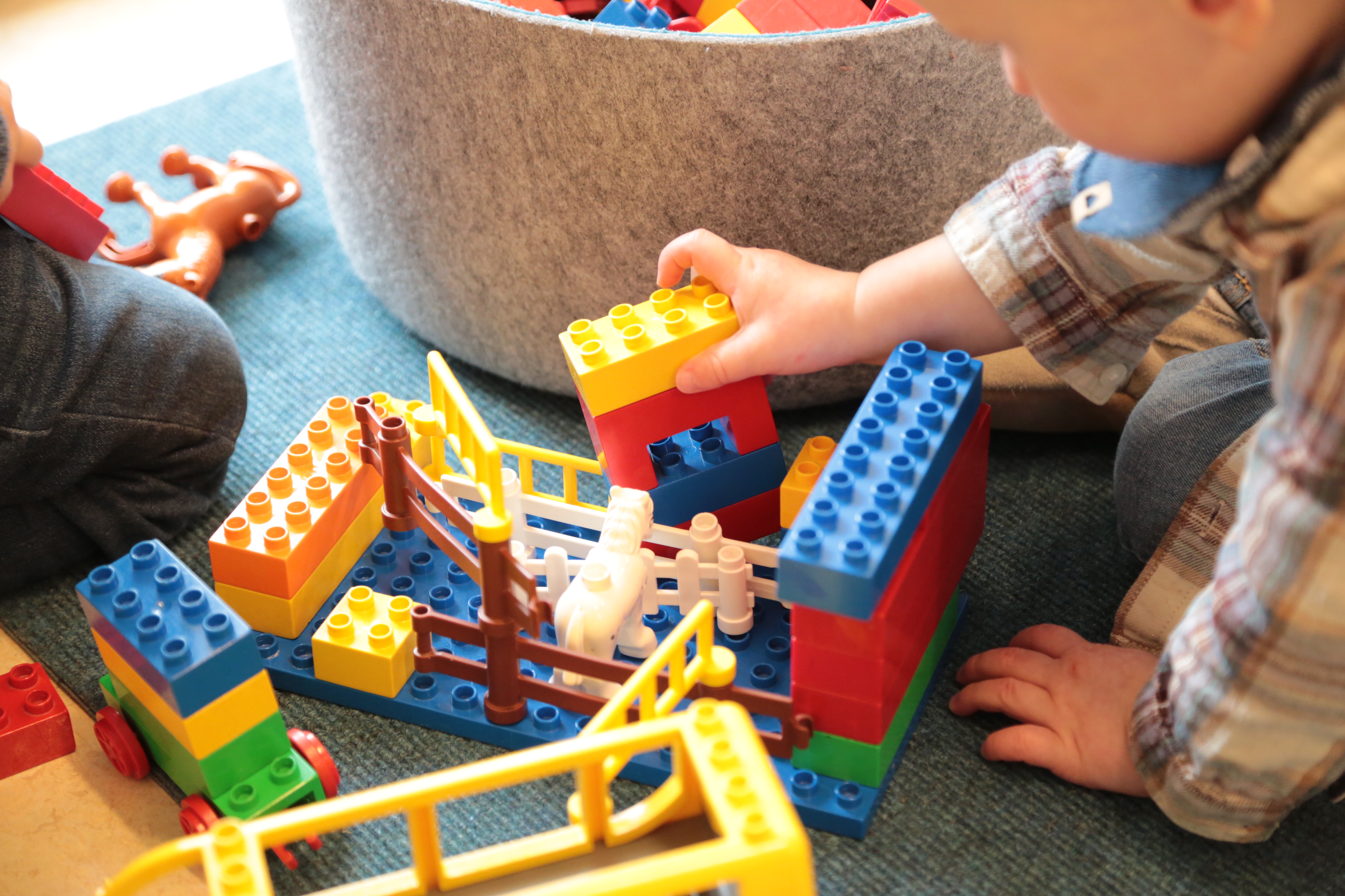 Von rechts greift ein kleines Kind nach Lego-Duplo-Steinen, ein zweites Kind kniet daneben, nur die Beine sind zu sehen.