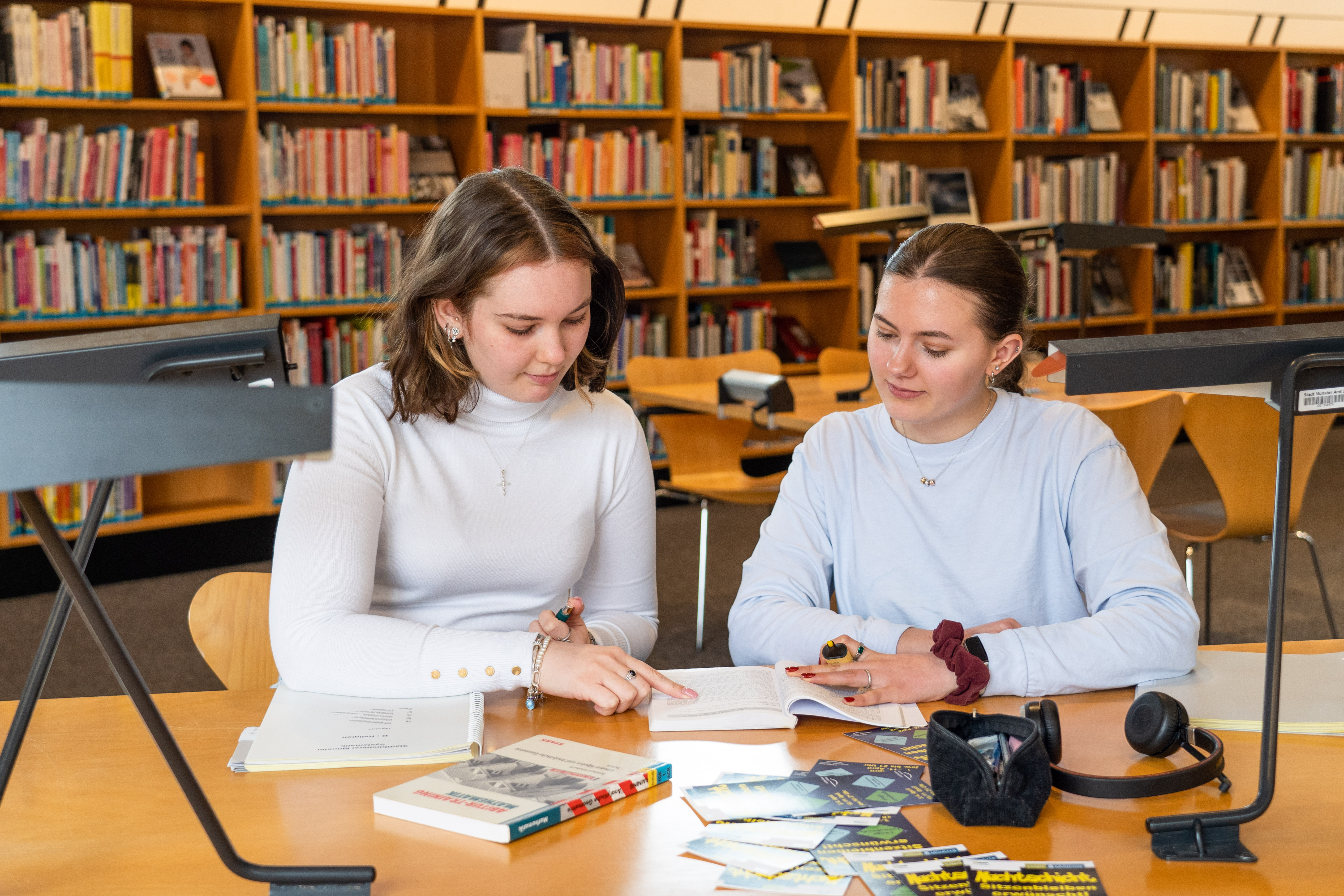 ZWei junge Mädchen sitzen an einem Tisch und schauen in ein Buch. Im Hintergrund stehen Bücherregale in der Bücherei.