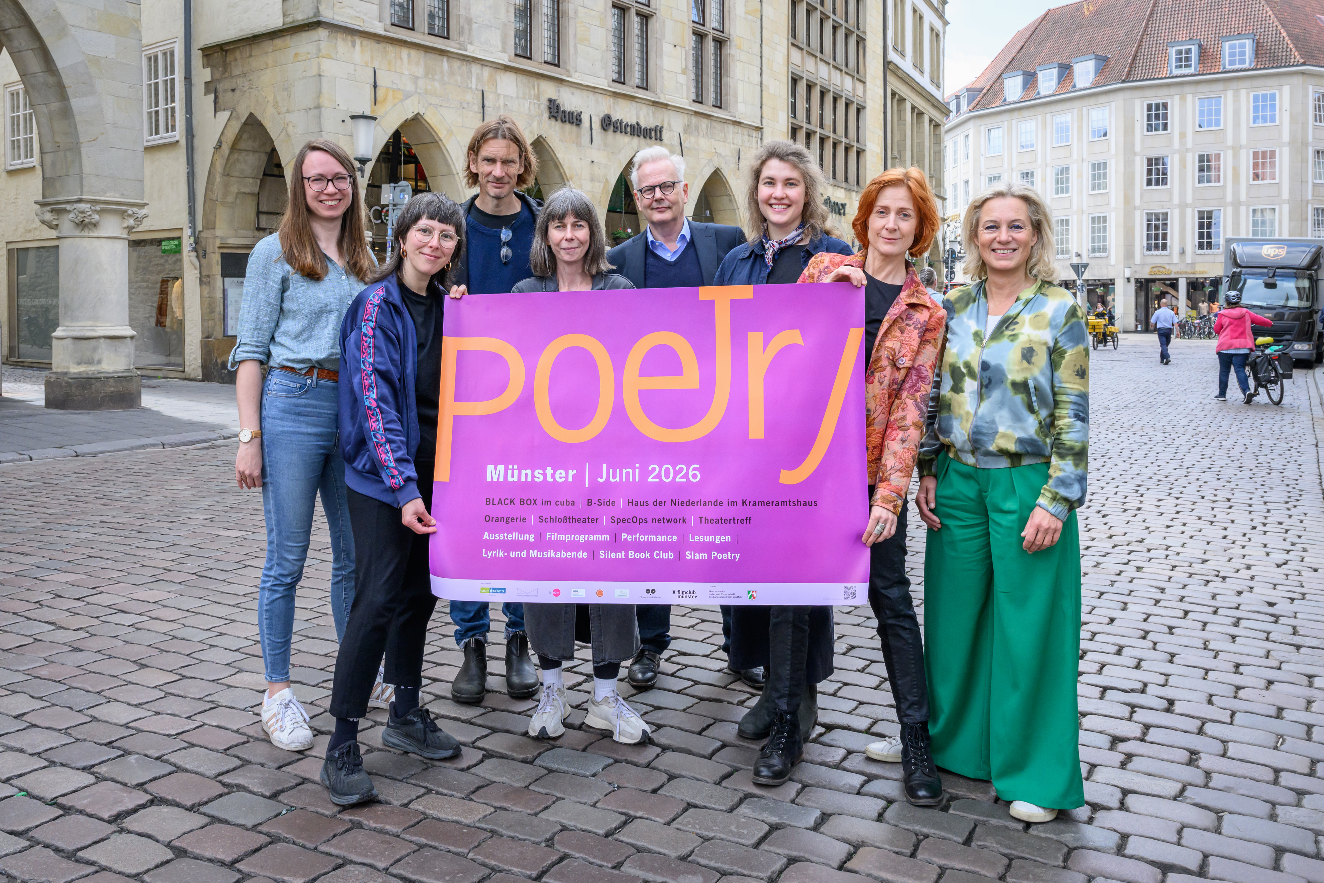 Organisatoren zeigen das aktuelle POETRY-Plakat auf dem Prinzipalmarkt in Münster.