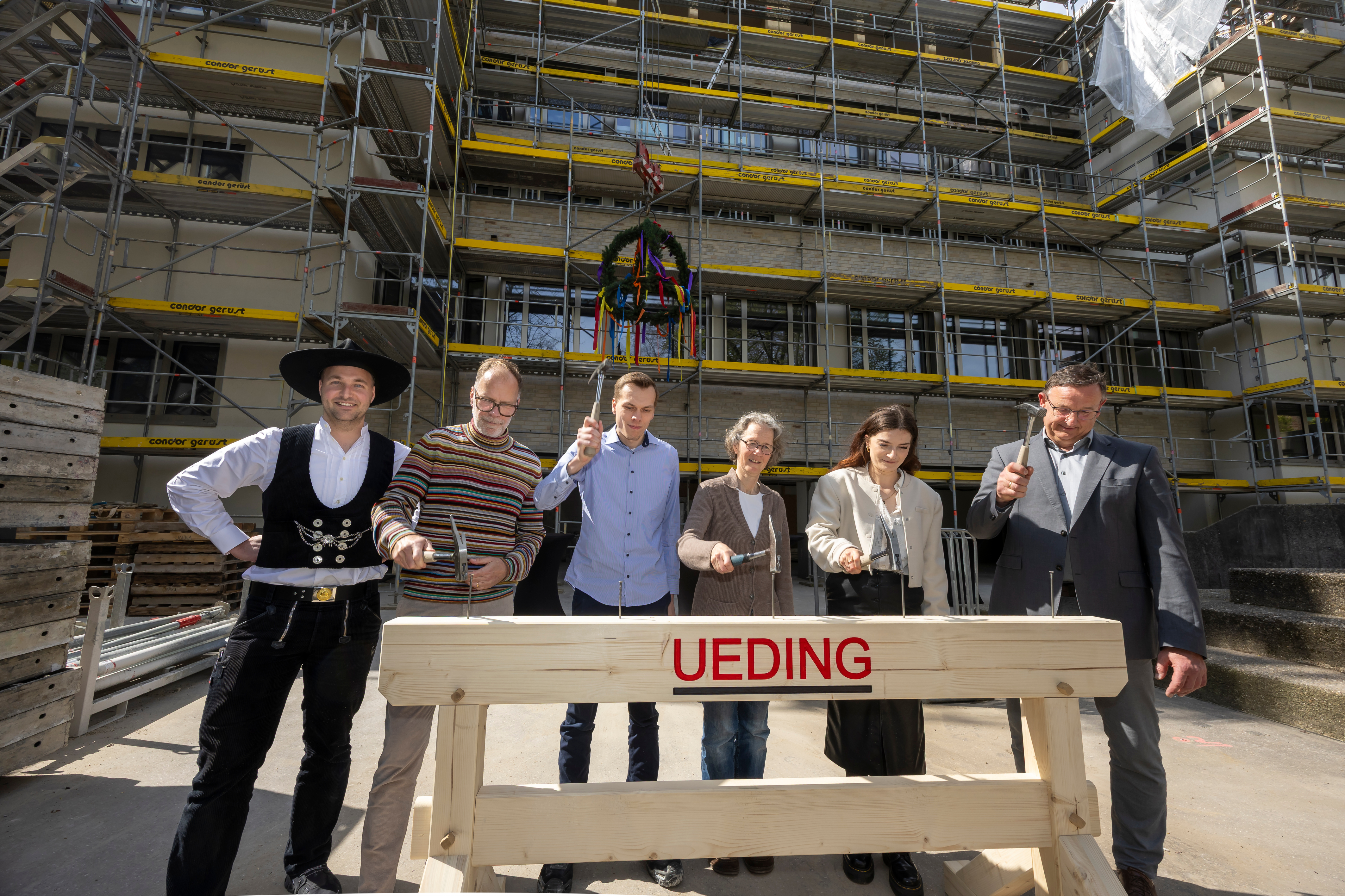 Sechs Personen schlagen jeweils mit einem Hammer auf einen Nagel in einem Balken.