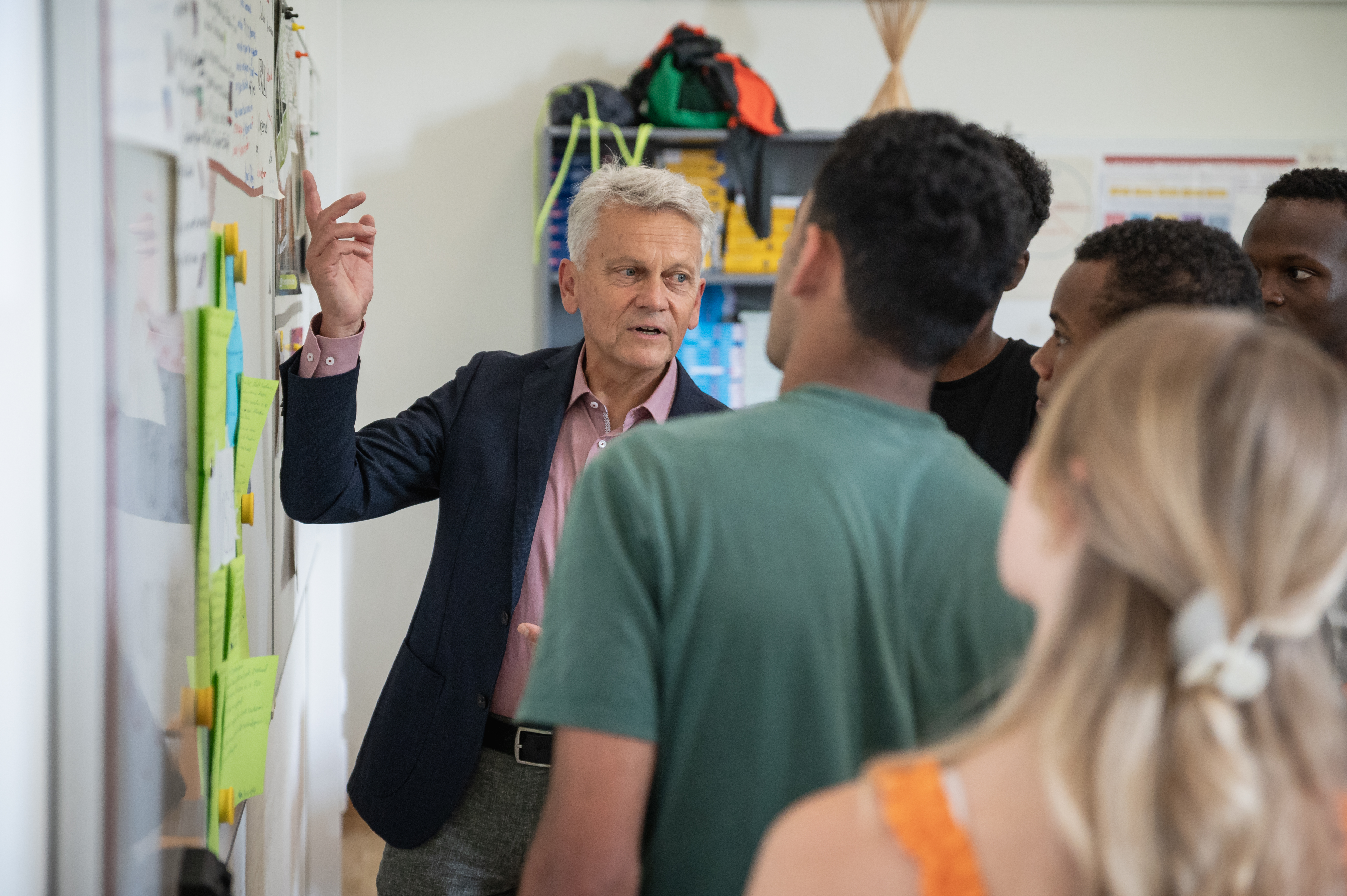 Ein Mann steht vor einer Gruppe junger Personen und zeigt dabei auf eine Tafel