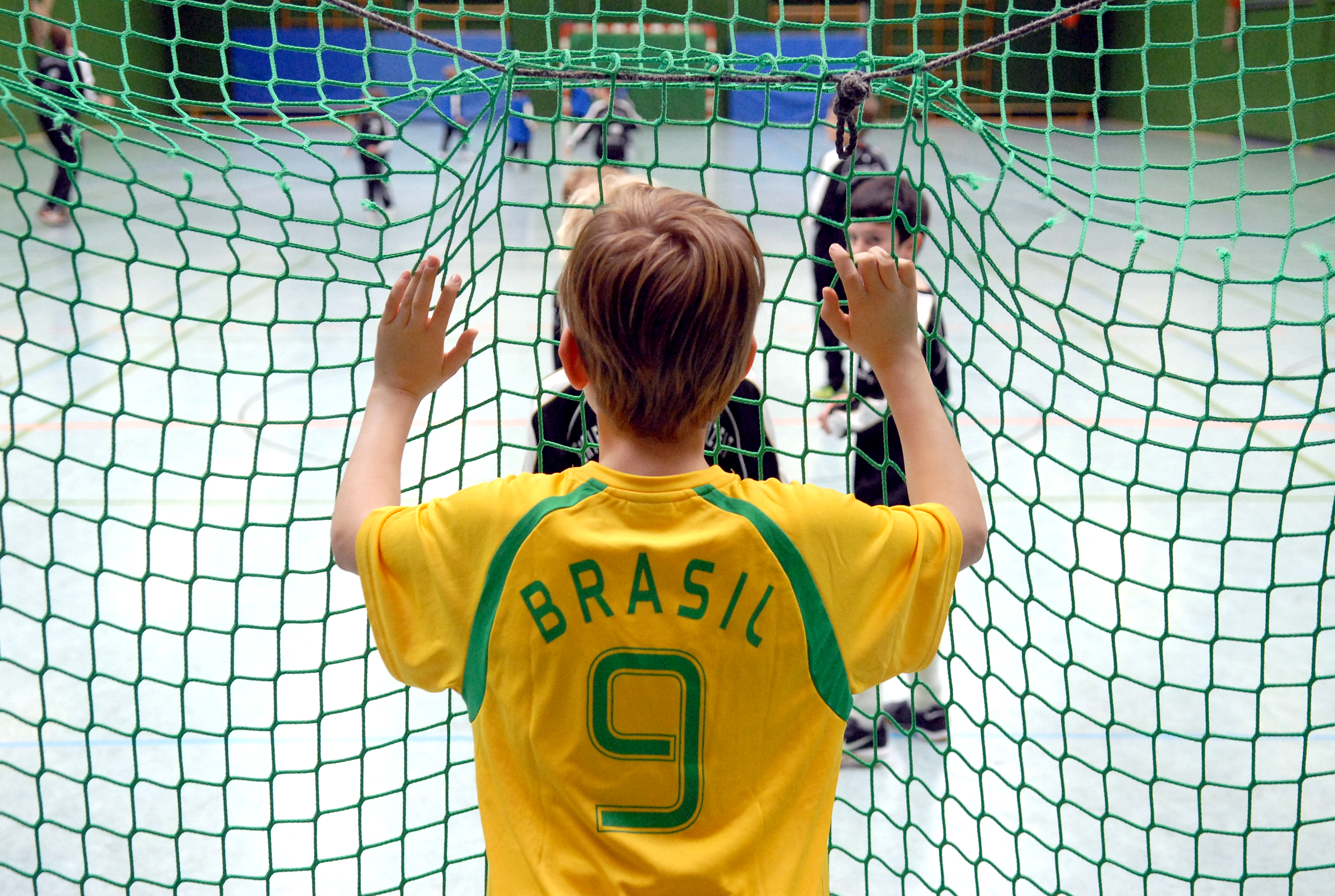 Kind mit Brasilien-Trikot steht hinter einem Tornetz und sieht anderen Kindern beim Fußball zu