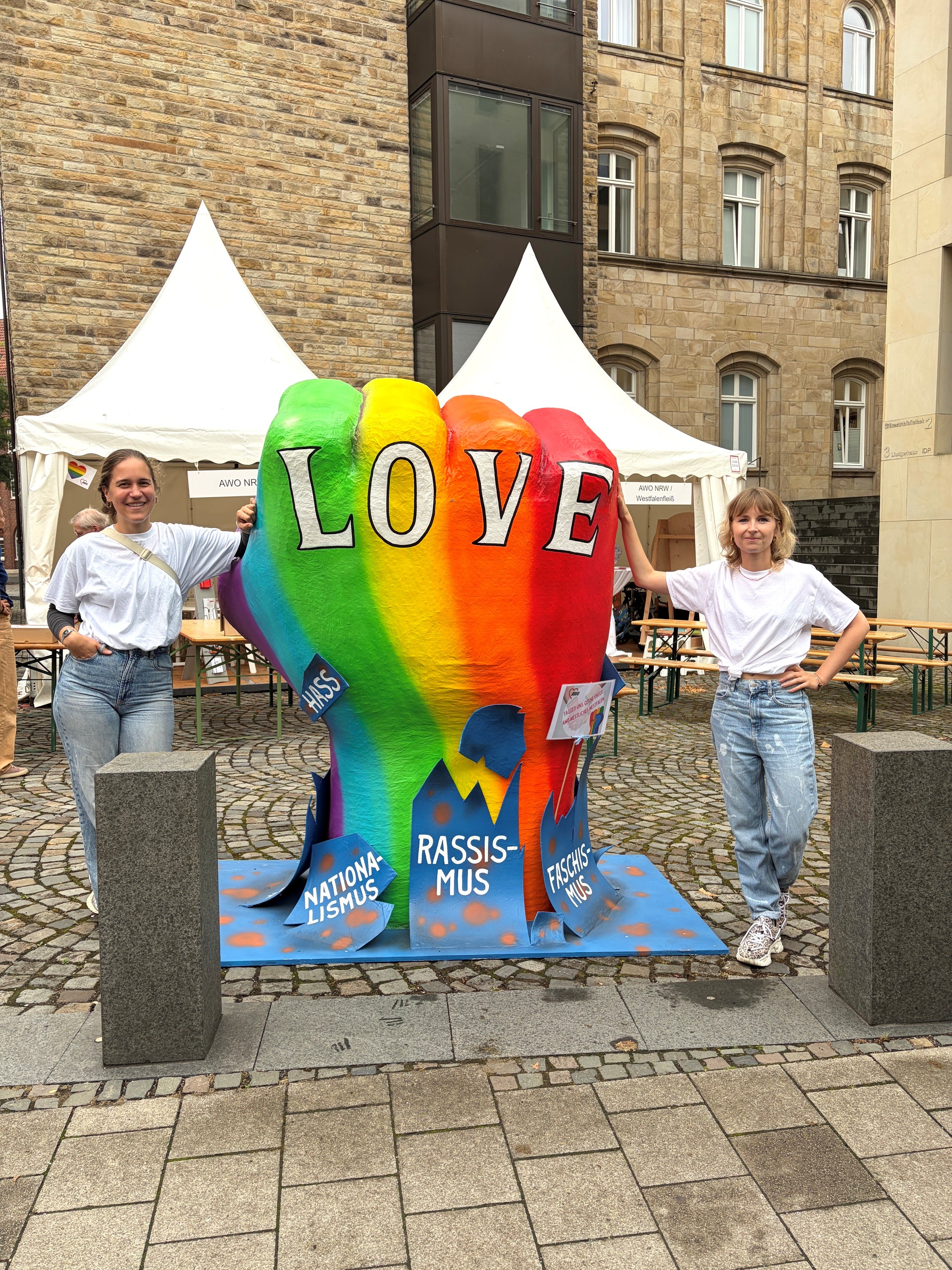 Zwei junge Frauen stehen neben einer Skultpur, die eine Faust darstellt, auf der groß das Wort "LOVE" steht. Die Faust hat die Farben des Regenbogens. Die Faust bricht symbolisch durch die Worte Nationalismus, Rassismus, Faschismus und Hass.