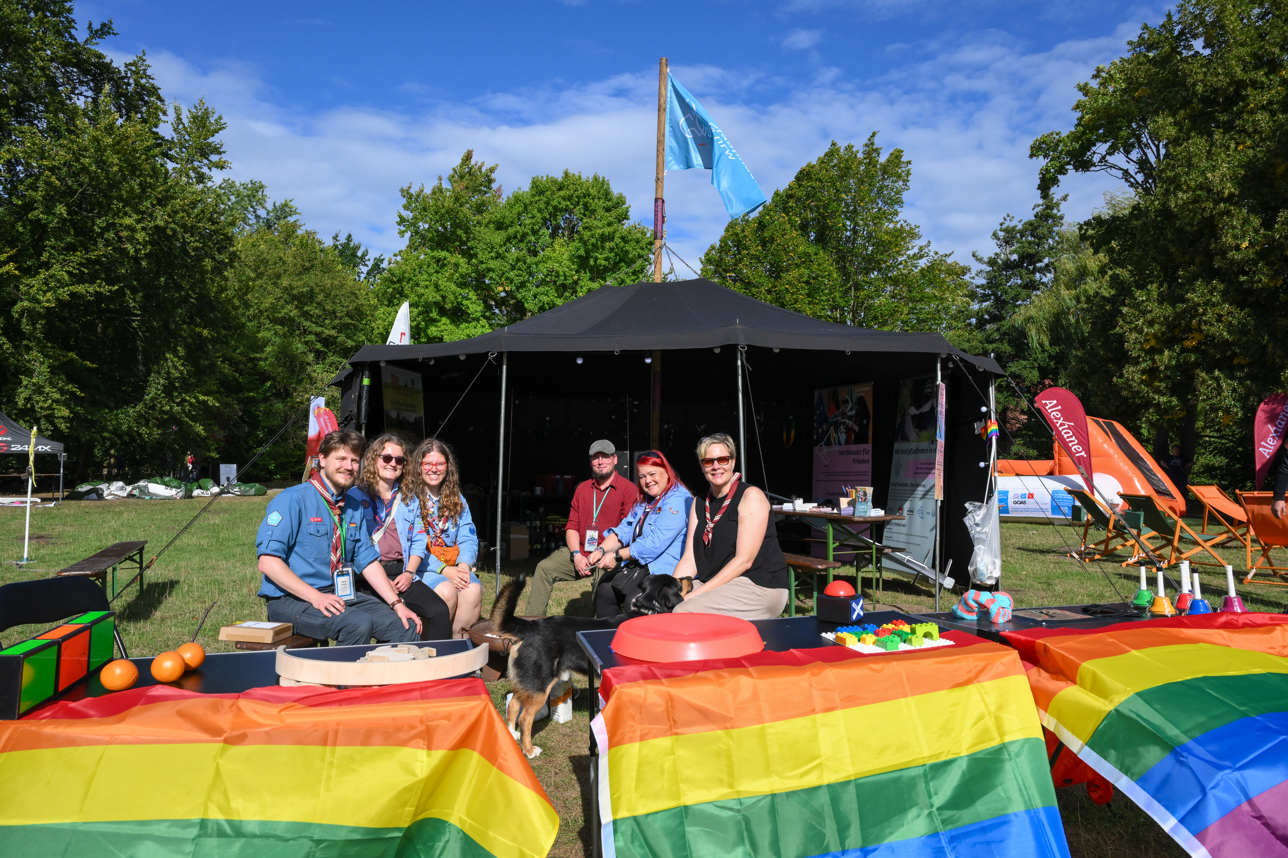 Einige Menschen sitzen vor einem Zelt. Es sind kleinere Stände aufgebaut. Man sieht Regenbogenflaggen und Spielzeug wie Bälle oder Lego