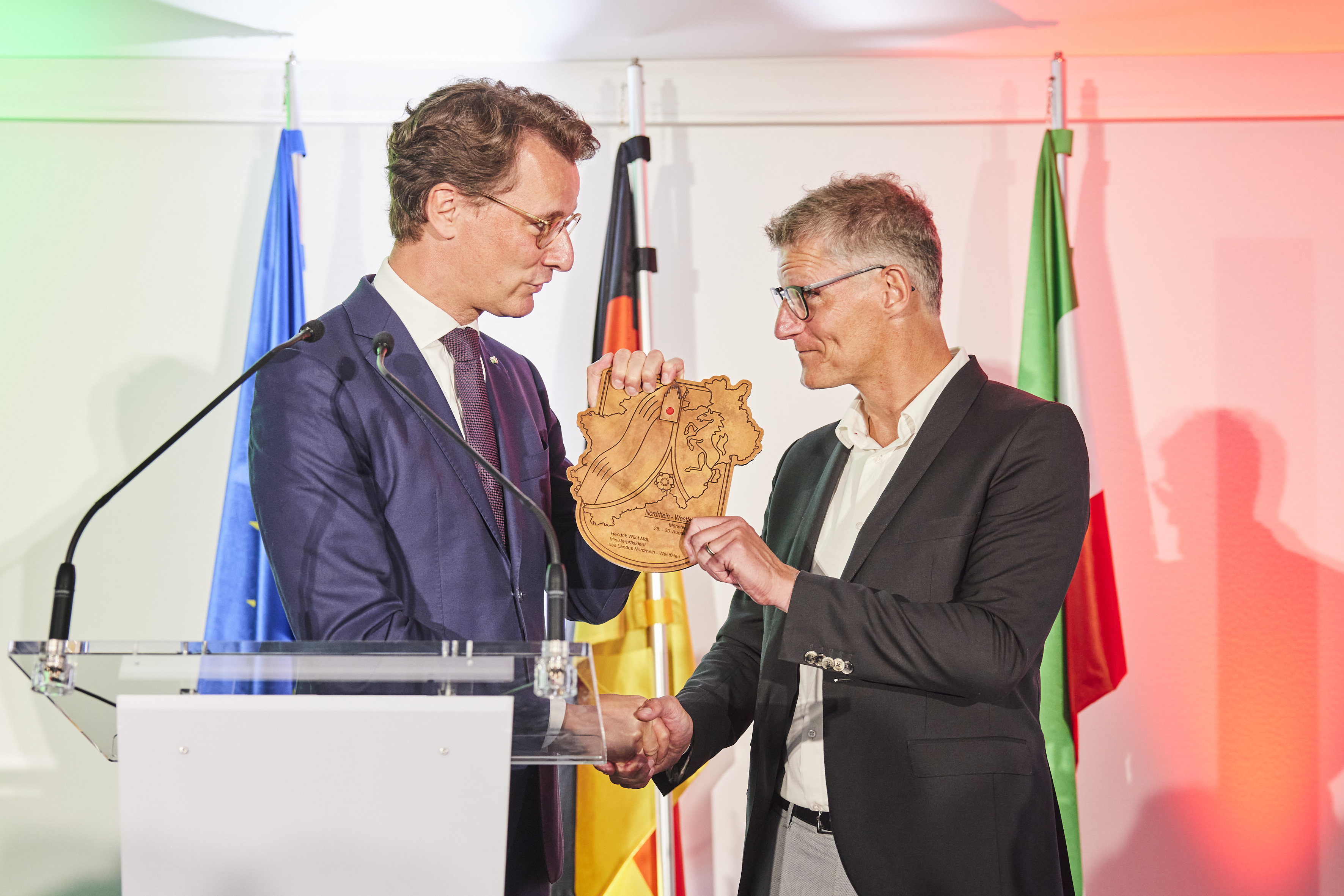 Ministerpräsident Hendrik Wüst überreicht Oberbürgermeister Tilman Fuchs einen Holzschnitt mit dem Umrissen von NRW.