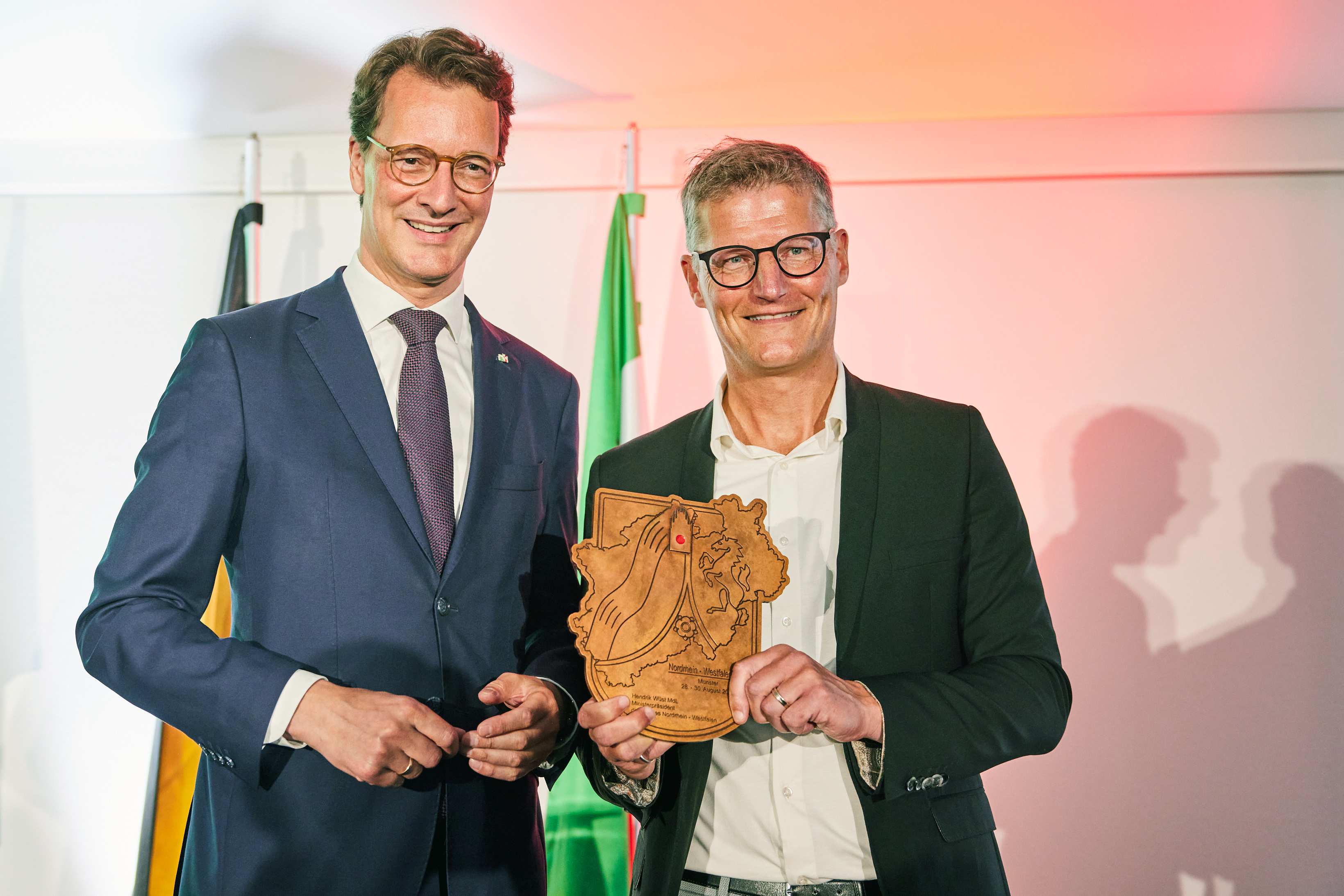 Hendrick Wüst und Timan Fuchs lächeln in die Kamera und halten eine Holztafel.