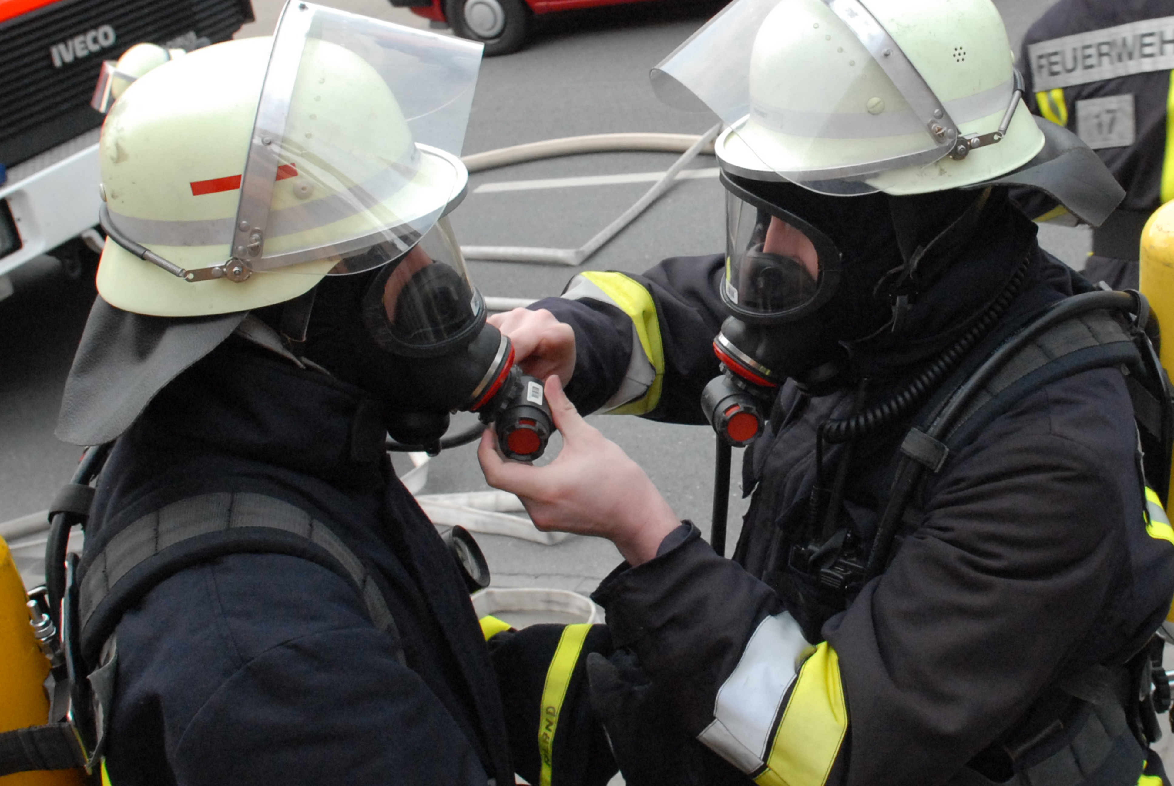 Zwei Feuerwehrmänner in voller Montur mit Atemmasken. Der Rechte hilft dem Linken beim Anlegen der Maske.