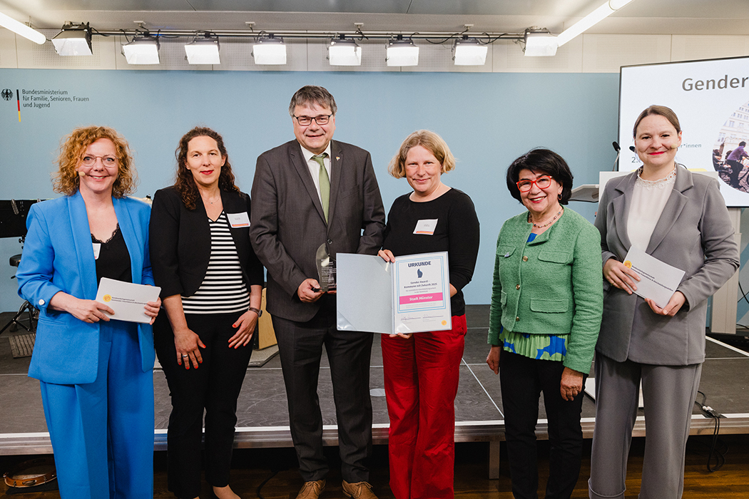Die Personen stehen aufgereiht vor einer Bühne des Bundesministeriums. Klaus Rosenau und Sarah Braun in der Mitte halten die Auszeichnung in Form eines gläsernen Pokals und einer Urkunde in den Händen.