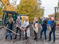 Gruppenbild: sechs Personen vor einem Bagger werfen mit Schaufeln Schotter in die Luft