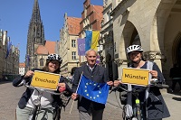 Mitarbeitende des Büros Internationales der Stadt Münster stehen auf dem Prinzipalmarkt mit ihren Fahrrädern. Sie halten gelbe Ortsschilder 'Münster' und 'Enschede' sowie eine Europafahne in den Händen.