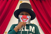 Ein Kind mit schwarzem Hut steht in einem geöffneten roten Vorhang und hält sich eine Karte mit einem lachenden Clownsmund vors Gesicht.