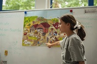 Mädchen zeigt auf ein Plakat auf dem ein Bauernhof abgebildet ist