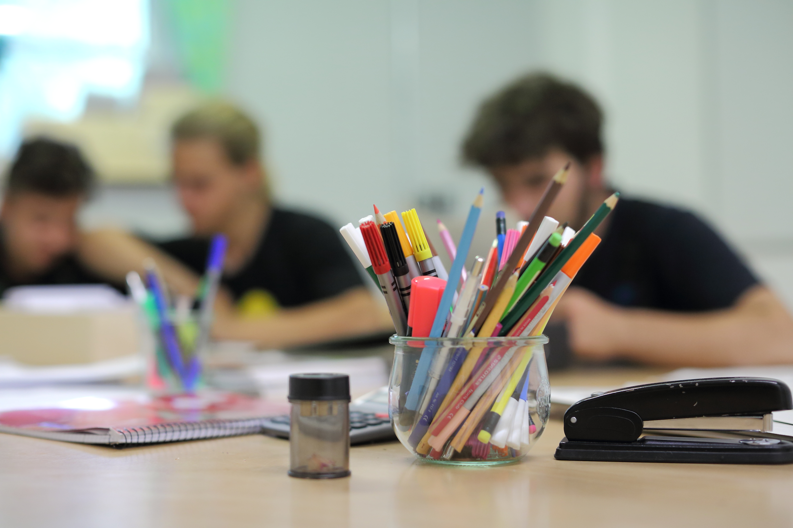 Unscharf zu erkennen arbeiten SchülerInnen im Hintergrund im Unterricht, im Vordergrund scharf zu sehen ist ein Glas mit Buntstiften.