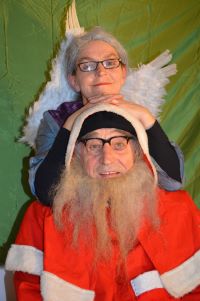 Foto: Frank Oelkers Eine ältere Frau mit Engelsflügeln stützt sich von hinten auf den Kopf eines älteren Mannes im Weihnachtsmannkostüm.