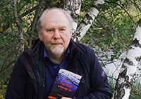 Porträtfoto von Georg Bühren, der mit seinem Buch in der Hand vor Birkenstämmen an einem Wasserlauf steht.
