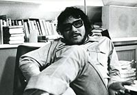Schwarz-weiß-Foto eines bärtigen dunkelhaarigen Mannes mit Brille, der mit übereinandergeschlagenen Beinen in einem Sessel lehnt.