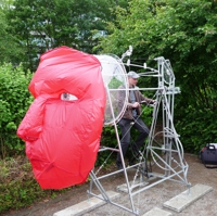 Großes rotes Stoffgesicht, das durch einen Popeller aufgeblasen wird, der durch ein Fahrrad angetrieben wird.