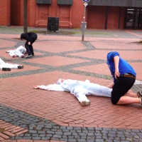 Foto von Jugendlichen, die sich über zwei Jugendliche in weißen Schutzanzügen beugen, die auf dem Boden liegen