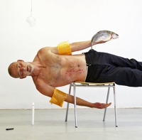 Bild zur Performance: Mann seitwärts auf einem Stuhl liegend, mit Schwimmflügeln an Oberarmen und Fußgelenken und mit einem Fisch in der linken Hand.