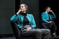 Zwei Frauen, eine ältere und eine jüngere, sitzen gleich gekleidet auf einem Stuhl hintereinander auf der Bühne.