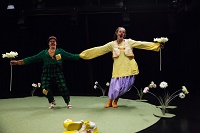 Ein Clown und eine Clownin halten sich an ausgestreckten Händen. In der anderen Hand halten sie jeweils eine große weiße Blume.
