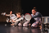 Eine Tänzerin und zwei Tänzer hocken auf dem Bühnenboden und strecken jeweils mit jedem Arm einen Topfdeckel vor sich aus.