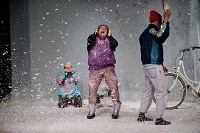 Die drei Performer*innen der Vorstellung stehen und hocken verteilt auf der Bühne und reagieren ganz unterschiedlich auf einen Schneefall aus Papier.