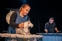 Im Vordergrund ist die Schauspielerin Anna Murböck zu sehen. Sie hält die Hauptfigur, eine Ziege, in der linken Hand. Im Hintergrund spielt der Musiker Jens Brülls auf dem Vibraphon.