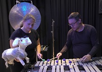 Im Vordergrund ist die Schauspielerin Anna Murböck zu sehen. Sie hält die Hauptfigur, eine Ziege, in der linken Hand. Im Hintergrund spielt der Musiker Jens Brülls auf dem Vibraphon.