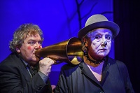 Ein älterer Schauspieler steht hinter einer älteren Schauspielerin und hält ein goldenes Sprachrohr an ihr Ohr. Beide tragen einen schwarzen Hut und rote Punkte auf Nase und Wangen.