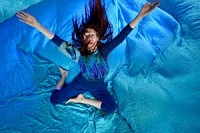 Die Tänzerin Dagmar Chittka sitzt mit angewinkelten Beinen und ausgestreckten Armen in tänzerischer Haltung auf einem hellblauen Seidentuch.