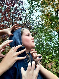 Eine Schauspielerin des Theaterkinderclubs steht mit aufgezogener Kapuze im seitlichen Profil. Fünf Hände greifen nach ihrem Kopf.