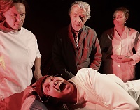 Drei Akteure, zwei davon im weißem Kittel, stehen beobachtend hinter einem "Patienten". Dieser liegt mit weit aufgerissenem Mund und schmerzverzerrtem Gesicht auf einer Bahre.