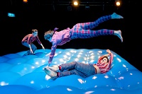 Ein Schauspieler und zwei Schauspielerinnen hüpfen auf einem bühnenfüllenden blauen Luftkissen.