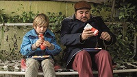 Ein Junge sitzt neben einem Mann mit Mütze auf einem Matratzengestell. Beide halten eine Ketchupflasche in Händen und spritzen Ketchup auf ihr Pommes frites.