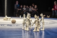 Auf der Bühne sind kleine Figuren, kreiert aus Kreppklebeband, kreisförmig aufgestellt. Im Hintergrund ist das junge Publikum mit Begleitpersonen schemenhaft zu erkennen.