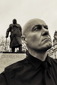 Im Hintergrund ist eine Statue von Winston Churchill zu sehen. Der Schauspieler Tom Corradini Teatro steht im seitlichen Profil davor und schaut ernst.