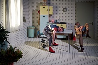 Der Bühnenort ist eine Küche. Die Schauspielerin liegt mit ihrem Oberkörper und geschlossenen Augen auf dem Tisch. Der Schauspieler hat ein Bein über einen Stuhl gelegt und sitzt mit gebeugtem Oberkörper nach hinten auf Diesem.
